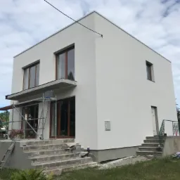 Dwukondygnacyjny dom w stylu nowoczesnej kostki, świeżo po malowaniu elewacji na biało, z widocznymi drewnianymi oknami, schodami wejściowymi w trakcie wykańczania oraz rusztowaniem przy balkonie.