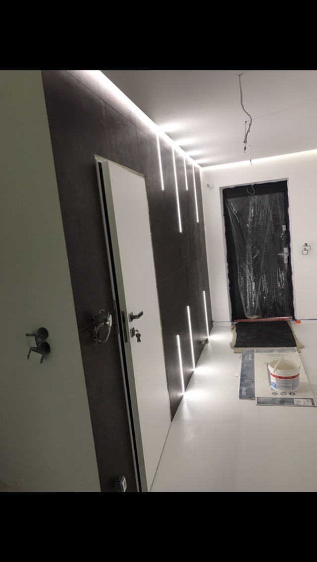 Wnętrze korytarza w trakcie prac wykończeniowych z zamontowanymi drzwiami, dekoracyjnym oświetleniem LED na ciemnej ścianie i zabezpieczonym wejściem do pomieszczenia folią ochronną.