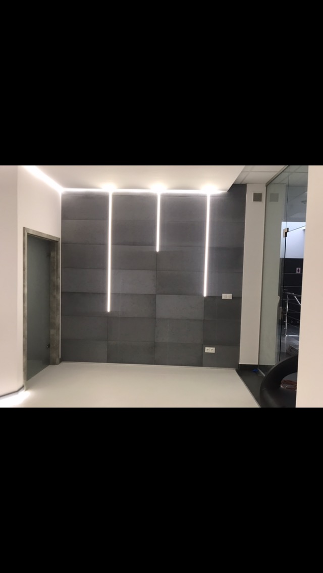Nowoczesna ściana z szarych paneli z trzema pionowymi liniami świetlnymi LED, białe ściany boczne i sufit z listwą LED, widoczne gniazdka elektryczne i ościeżnica drzwiowa.