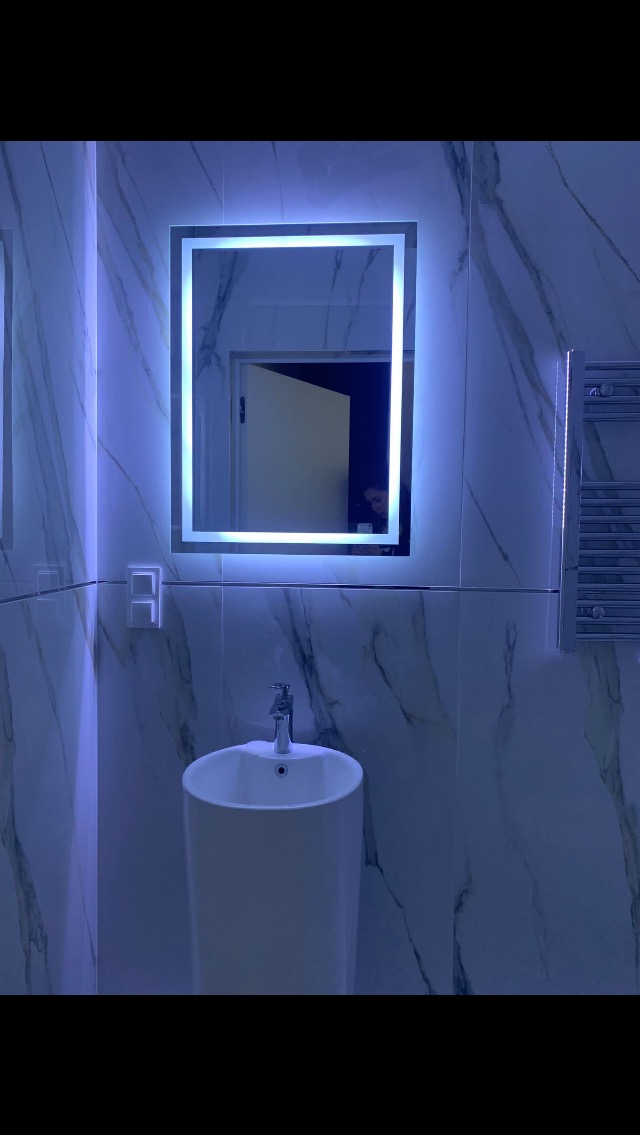 Nowoczesna łazienka z umywalką kolumnową i podświetlanym lustrem LED na marmurowej ścianie, widoczny grzejnik drabinkowy.