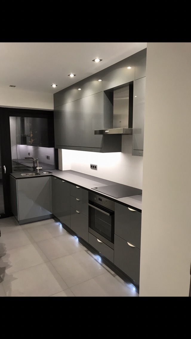 Nowoczesna kuchnia w odcieniach szarości z błyszczącymi frontami, oświetleniem podszafkowym i punktowym oraz płytkami podłogowymi w jasnym odcieniu szarości.