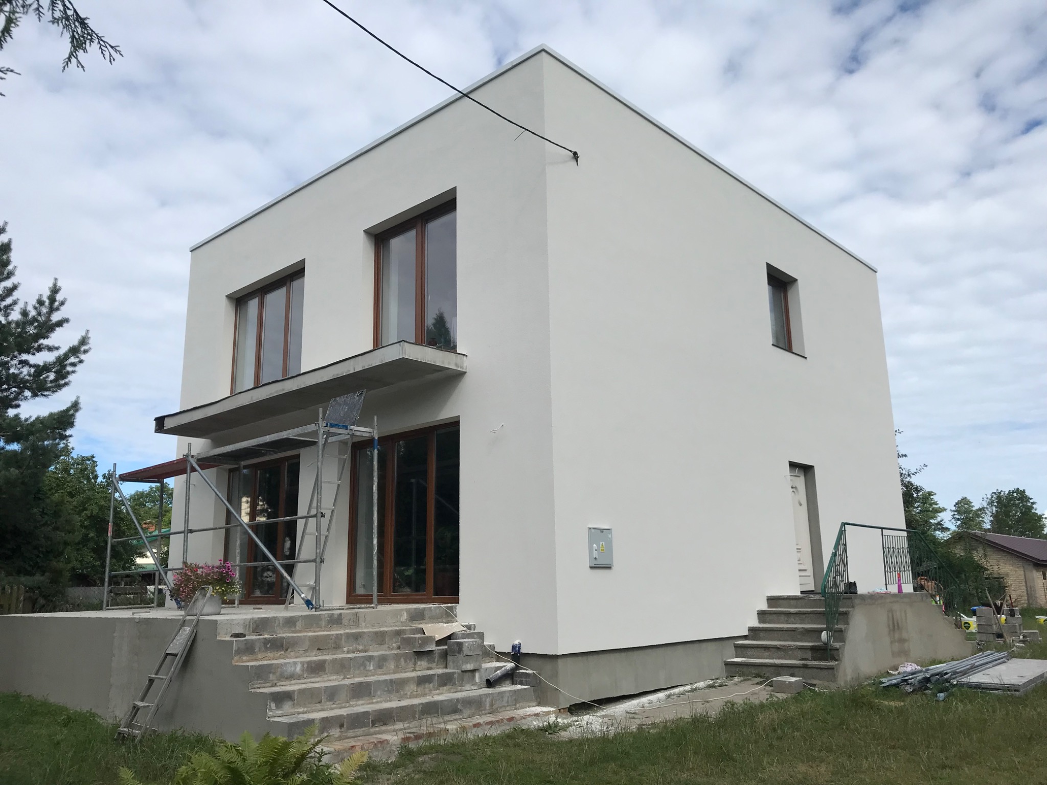 Dwukondygnacyjny dom w stylu nowoczesnej kostki, świeżo po malowaniu elewacji na biało, z widocznymi drewnianymi oknami, schodami wejściowymi w trakcie wykańczania oraz rusztowaniem przy balkonie.
