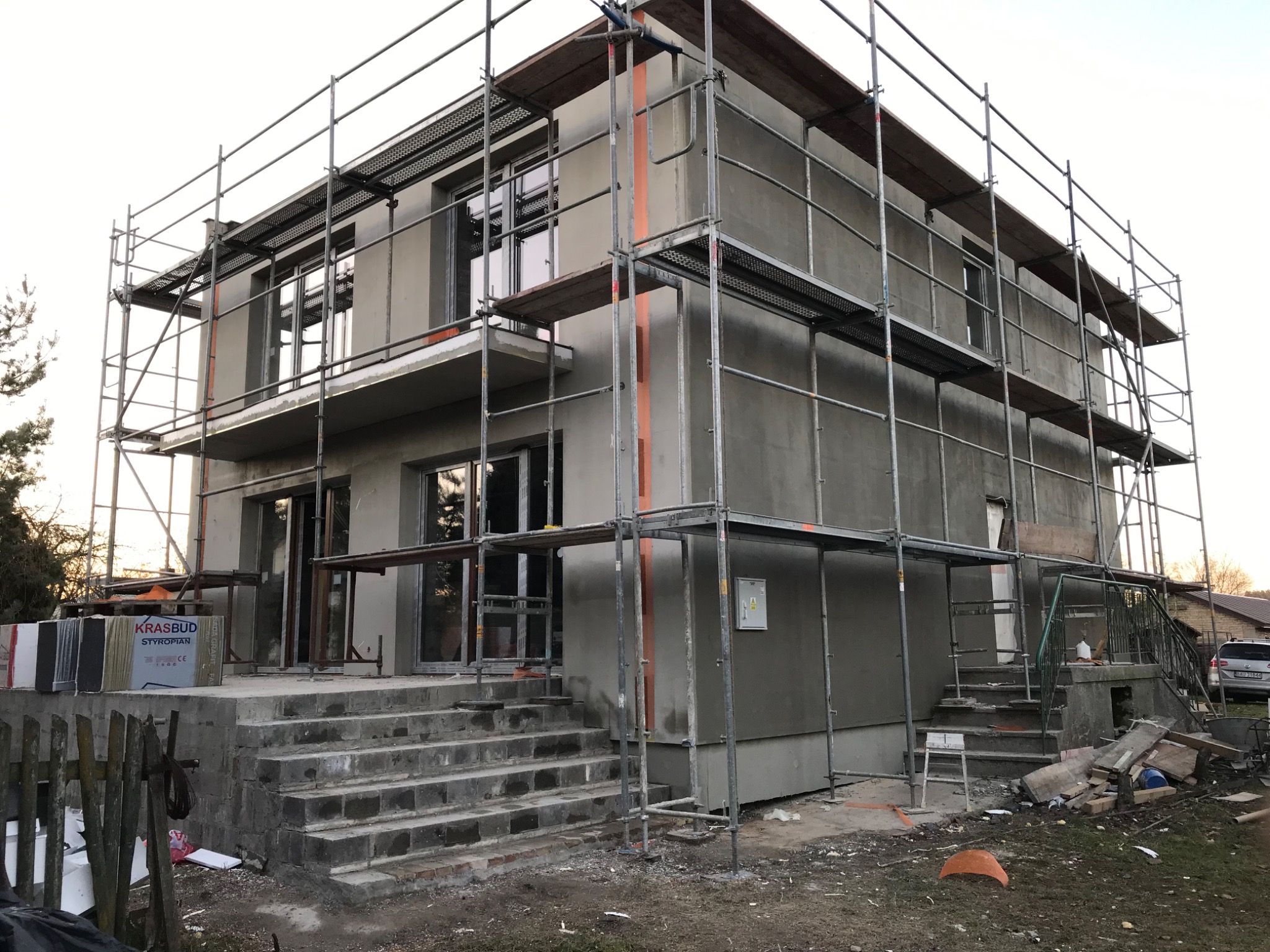 Surowy dom dwukondygnacyjny w trakcie ocieplania elewacji, otoczony rusztowaniem, z widocznymi paczkami styropianu Krasbud na schodach wejściowych.