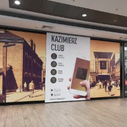 Duży baner reklamowy w galerii handlowej, promujący aplikację mobilną 'Kazimierz Club', z wizualizacją ulicy i logotypem galerii na ekranie smartfona.