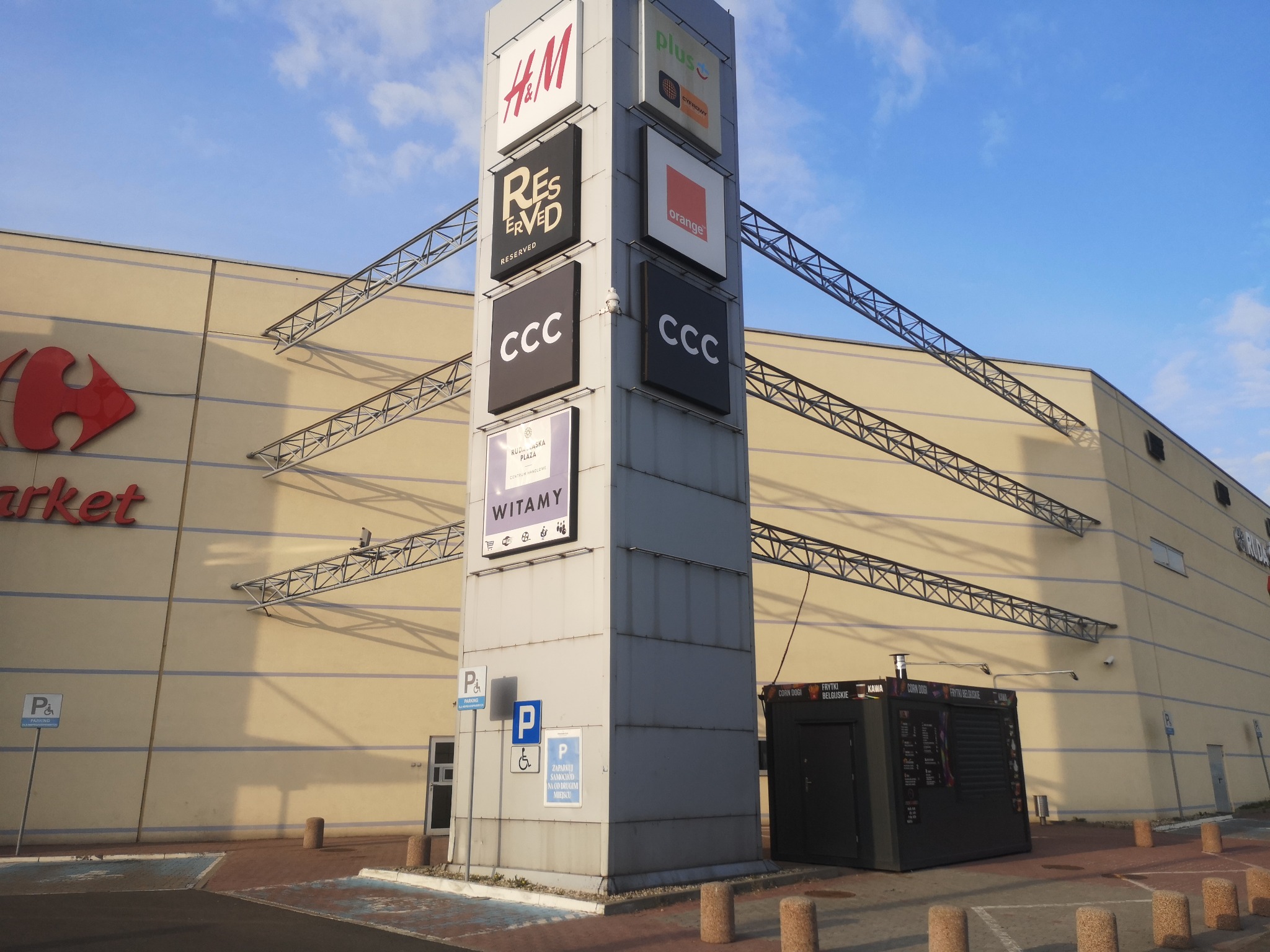 Pylon reklamowy z logotypami sklepów H&M, Reserved, CCC, Plus i Orange przed budynkiem galerii handlowej Ruda Śląska Plaza, widoczny także znak Carrefour Market.