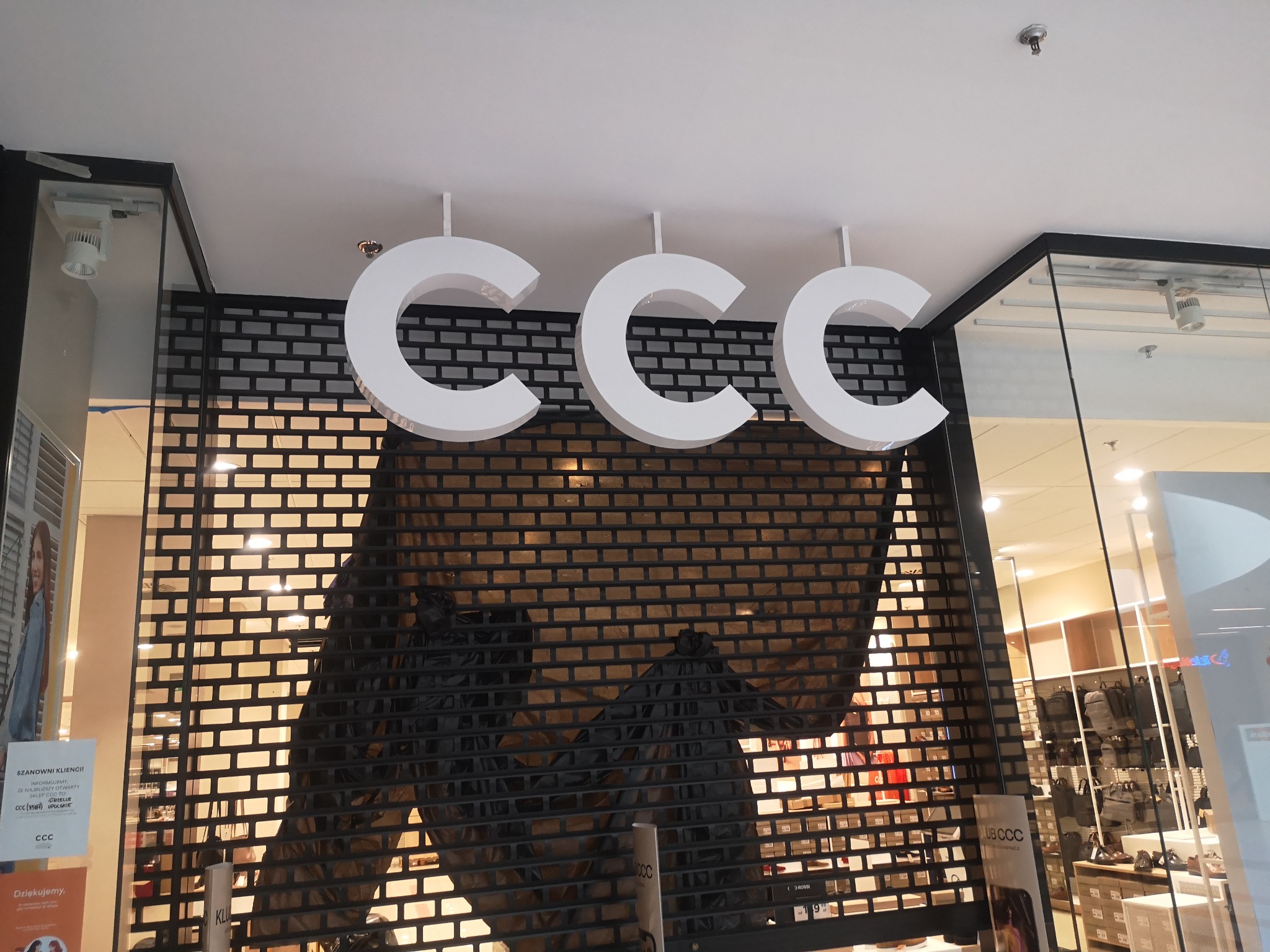 Trzy białe, podświetlane litery 'CCC' zawieszone pod sufitem nad zamkniętą, metalową roletą sklepu w galerii handlowej.