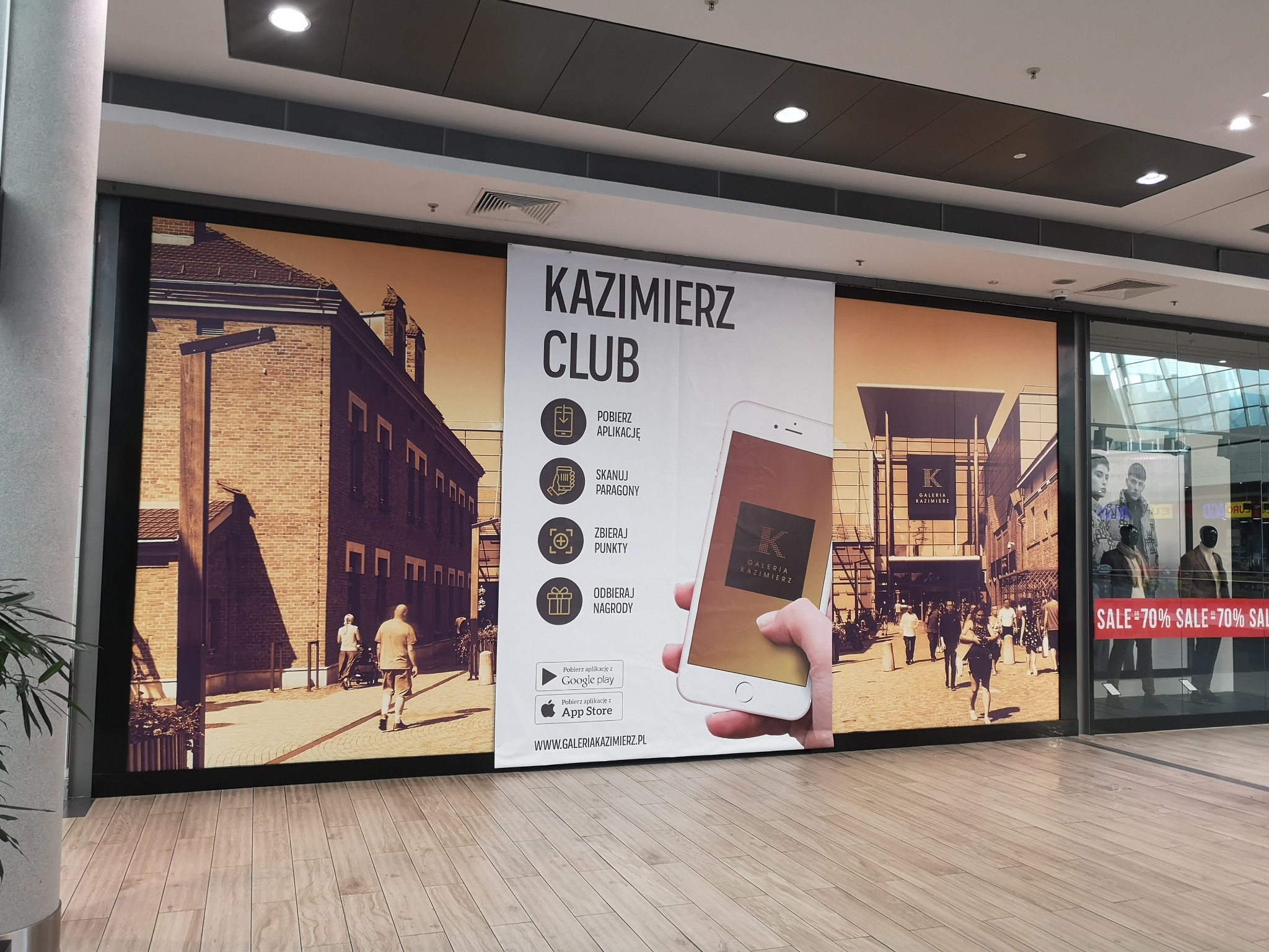 Duży baner reklamowy w galerii handlowej, promujący aplikację mobilną 'Kazimierz Club', z wizualizacją ulicy i logotypem galerii na ekranie smartfona.
