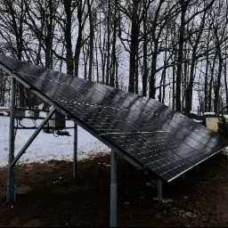 Panel słoneczny na metalowej konstrukcji, widoczny śnieg i drzewa w tle, pochmurny dzień.