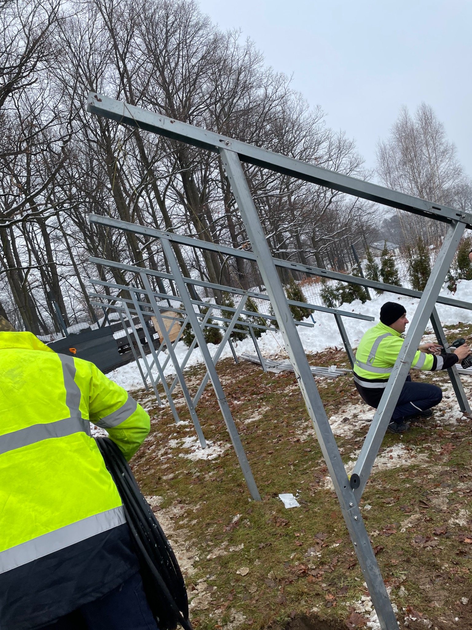 Montaż stalowej konstrukcji wsporczej pod panele na zewnątrz w zimowej scenerii. Dwóch monterów w odblaskowych kurtkach przykręca elementy konstrukcji. Na ziemi leży śnieg.