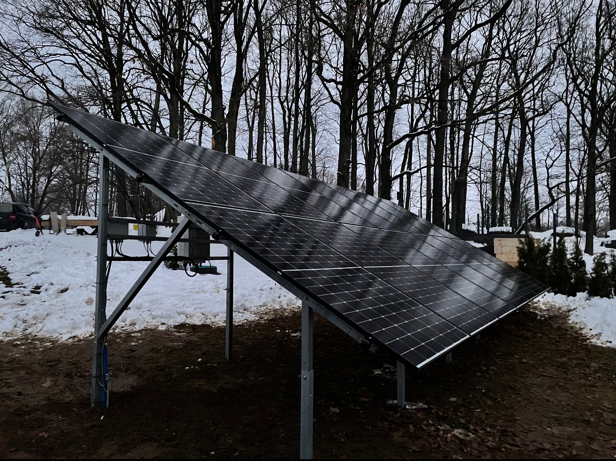 Panel słoneczny na metalowej konstrukcji, widoczny śnieg i drzewa w tle, pochmurny dzień.