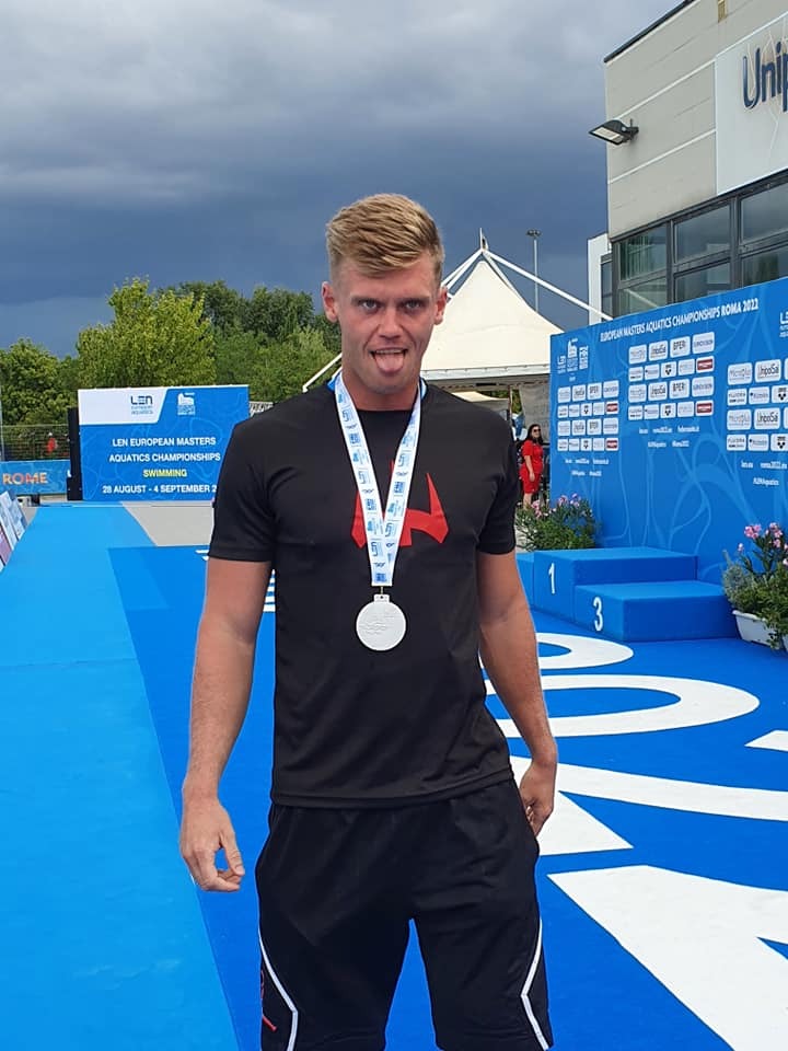 Uśmiechnięty pływak z blond włosami, z medalem na szyi, stojący na podium z numerami 1 i 3 w tle podczas zawodów LEN European Masters Aquatics Championships Roma 2022.