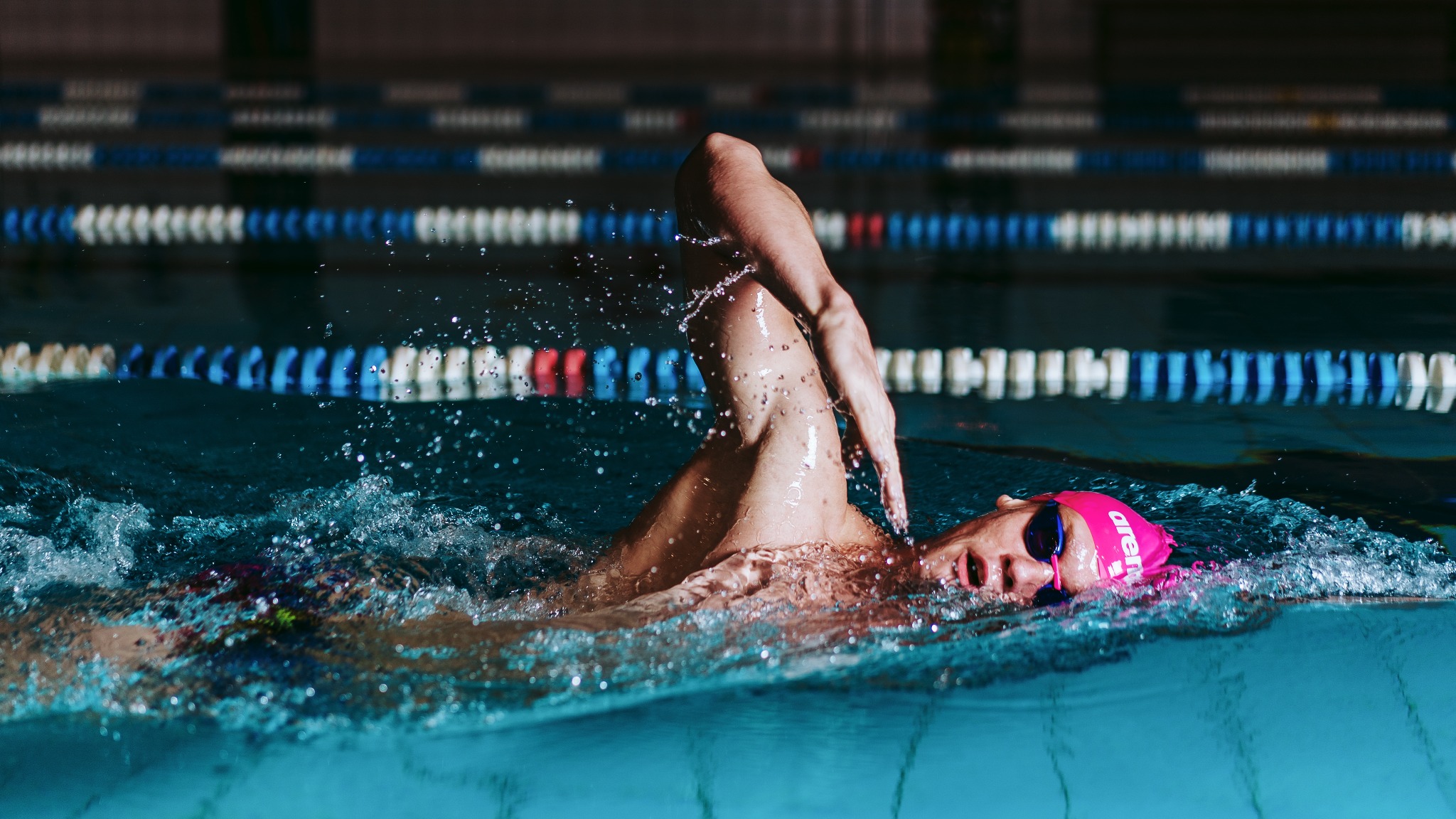 Pływak w różowym czepku Arena płynie stylem dowolnym, widoczne linie torów na basenie w tle.