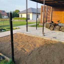 MAT-BRUK - Przygotowanie podłoża pod kostkę brukową na posesji z widoczną kostką, trawą, domami i mężczyzną w tle.