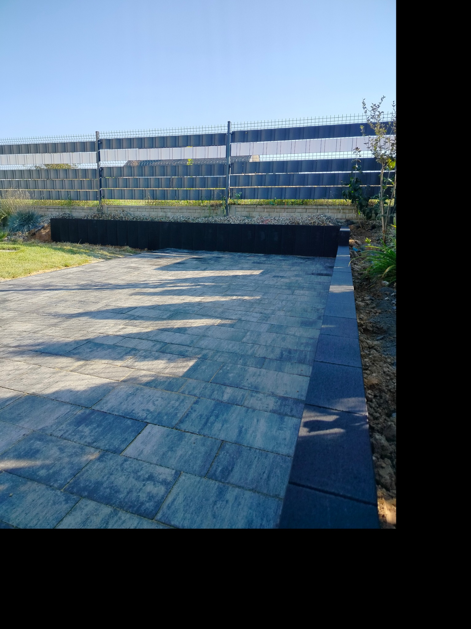Wykończony podjazd z kostki brukowej w odcieniach szarości i błękitu, obrzeże z ciemnej kostki, w tle panelowe ogrodzenie i rabata z roślinami, słoneczny dzień.