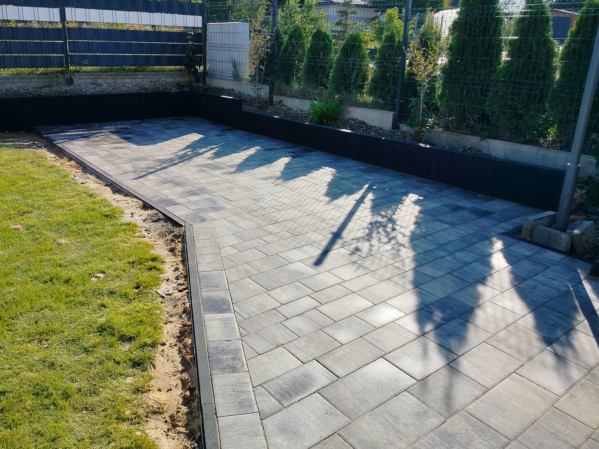 Nowo ułożona, szara kostka brukowa na podjeździe z widocznymi cieniami drzew, obrzeża z ciemnych kostek, trawnik po lewej stronie i czarne, betonowe donice w tle.