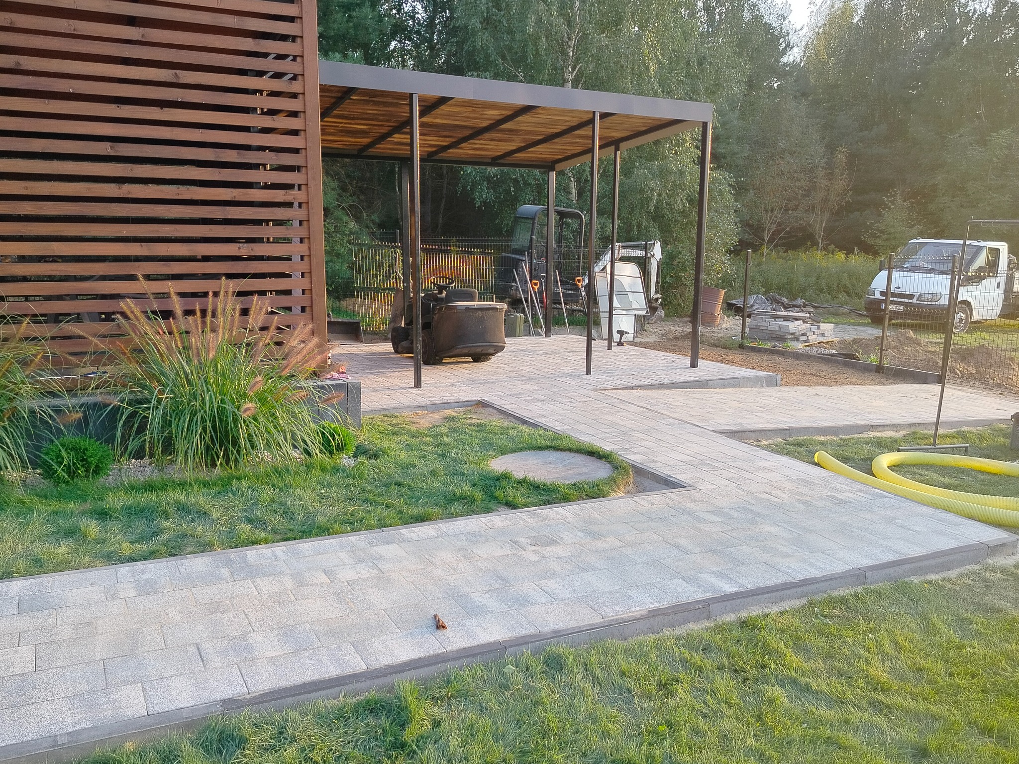 Ścieżka z jasnoszarej kostki brukowej prowadząca do zadaszonego miejsca postojowego z mini koparką w tle, obok drewniana ściana z ozdobną trawą i widoczny fragment ogrodzenia.