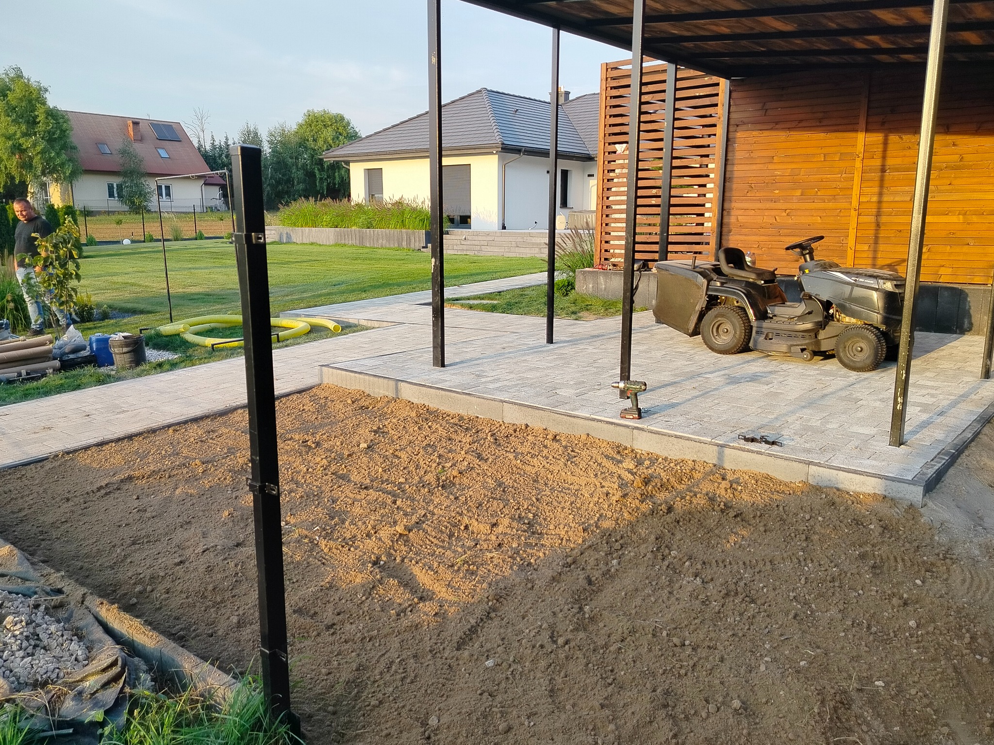 Przygotowanie podłoża pod kostkę brukową na posesji z widoczną kostką, trawą, domami i mężczyzną w tle.