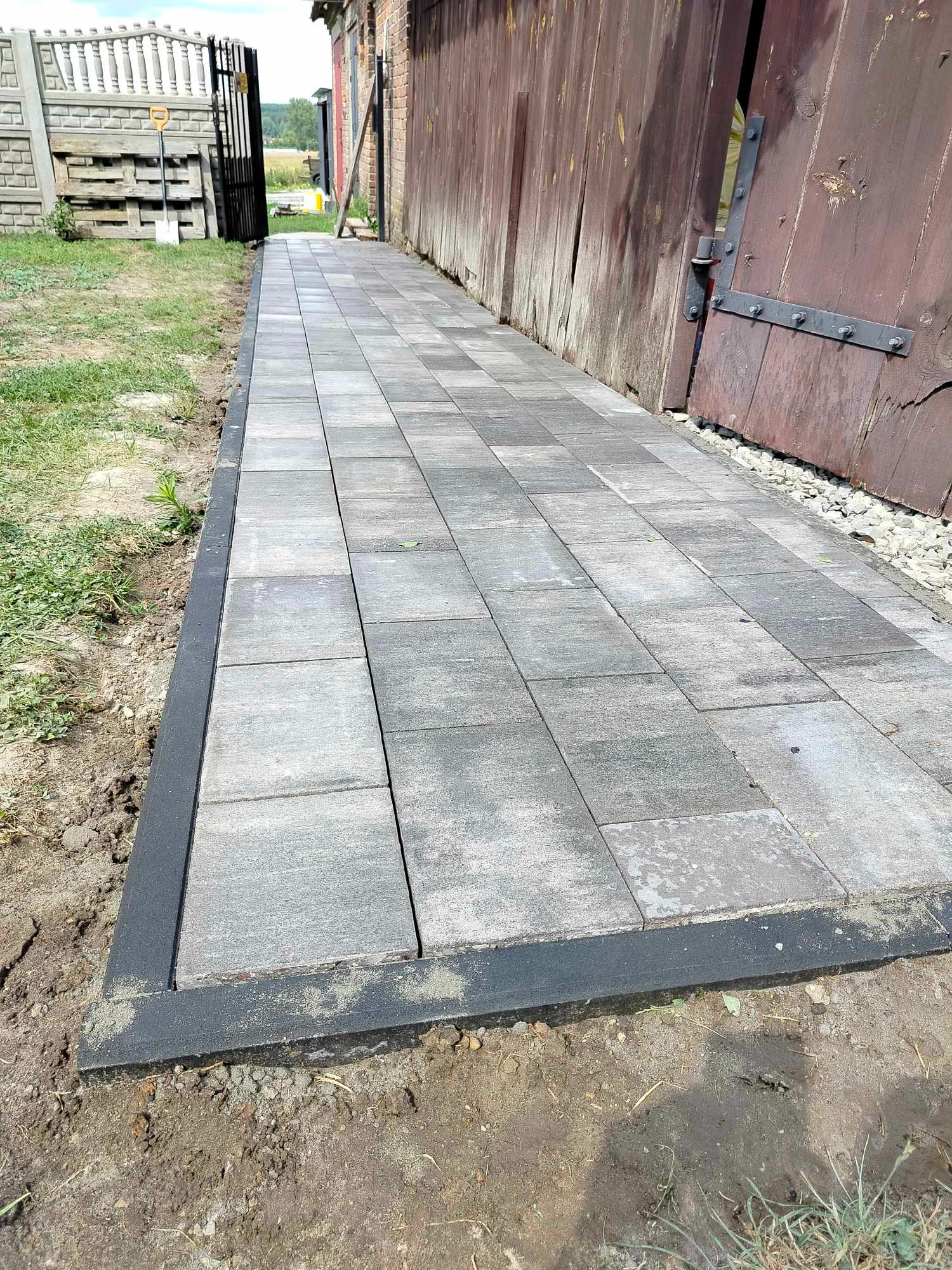 Ułożona ścieżka z szarej kostki brukowej z czarnym obrzeżem, prowadząca wzdłuż starej, drewnianej bramy w ogrodzie.