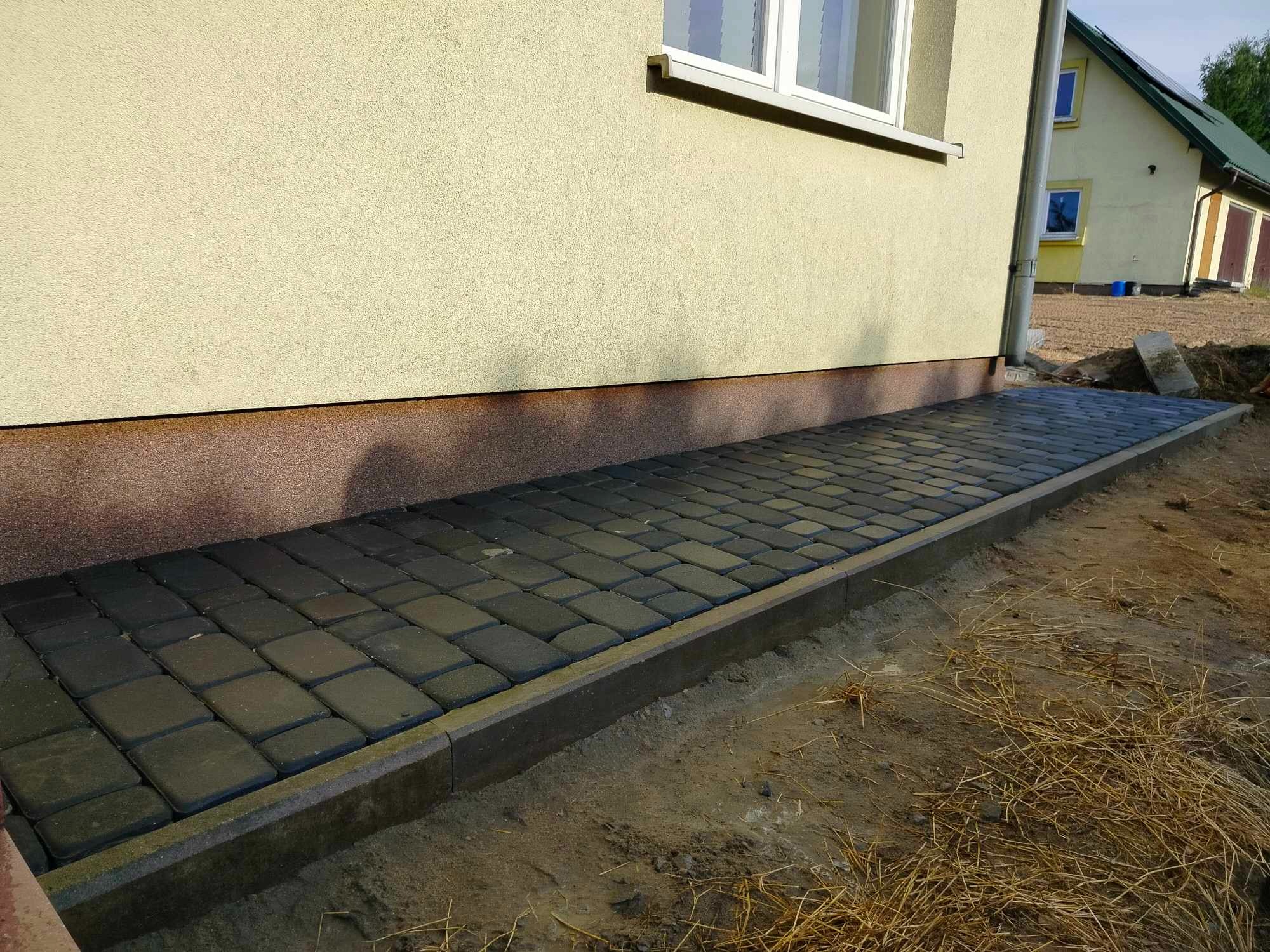 Nowo ułożona ścieżka z ciemnej kostki brukowej przy ścianie budynku z widocznymi elementami wykończeniowymi, otoczona ziemią i suchą trawą.