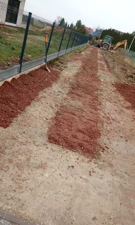 Nowo postawione ogrodzenie panelowe z podmurówką betonową, obok którego leży szpadel. W tle widoczny ciężki sprzęt budowlany przygotowujący teren pod drogę.