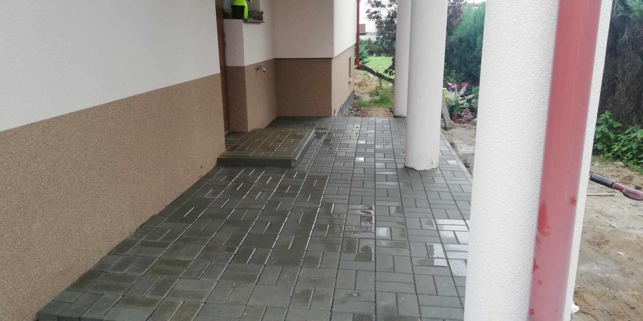 Świeżo ułożona, mokra kostka brukowa w odcieniach szarości tworząca chodnik przed wejściem do domu z kolumnami.