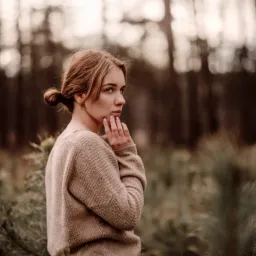 Portret młodej kobiety z włosami upiętymi w kok, w beżowym swetrze, z ręką przy twarzy, spoglądającej w bok na tle zamazanego leśnego krajobrazu.