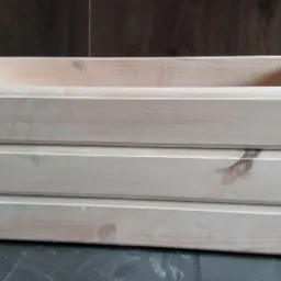 Surowy, drewniany kwietnik z jasnego drewna, wykonany przez stolarza z Lwówka Śląskiego, składający się z trzech poziomów desek, widoczne łączenia na rogach.