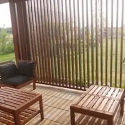 Zadaszony taras z drewnianą przesłoną z pionowych listew, brązowe meble ogrodowe: dwa stoliki kawowe i fotel z czarną poduszką, widok na ogród w tle.