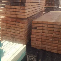 Stosy świeżo przetartego drewna ułożone na placu, zabezpieczone taśmą, z widocznymi śladami obróbki i naturalnym usłojeniem. W tle widoczne inne stosy drewna i elementy konstrukcji.