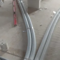 Wnętrze w trakcie budowy: rury instalacyjne w białej karbowanej osłonie, zamocowane metalowymi klamrami, ułożone równolegle na podłodze w surowym stanie, obok poziomica i niebieskie rury.