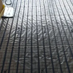 Montaż ogrzewania podłogowego: rury grzewcze marki Keller ułożone na srebrnej folii z nadrukowaną siatką, widok z góry w kierunku okna z rozproszonym światłem dziennym.