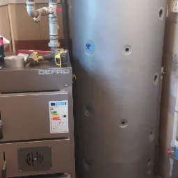 Kotłownia z kotłem DEFRO o mocy 17kW, zasobnikiem ciepłej wody i pompą obiegową na tle białych rur i brudnej podłogi z płytek.