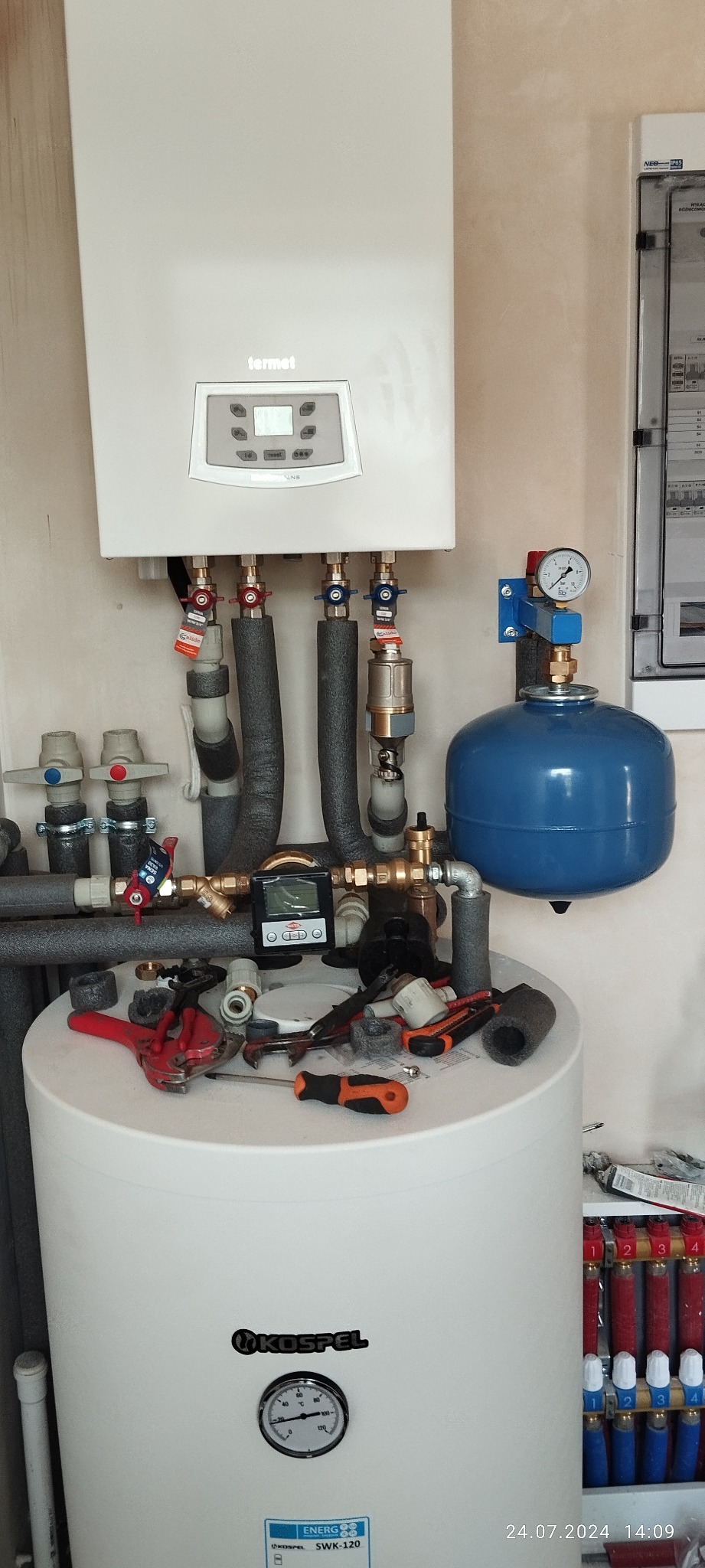 Montaż kotła gazowego Termet z widocznymi rurami, zaworami, manometrem i niebieskim zbiornikiem wyrównawczym; na górze zbiornika leżą narzędzia hydrauliczne, w tle skrzynka elektryczna.