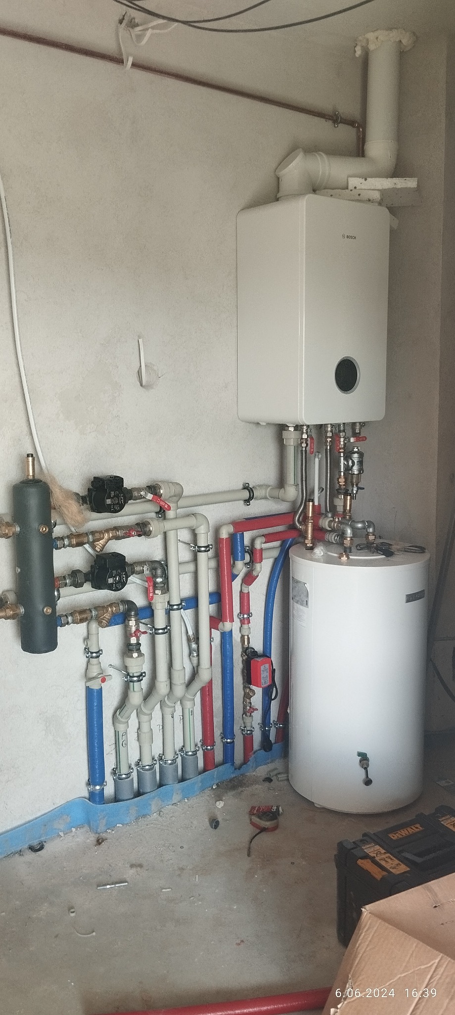 Nowoczesny kocioł gazowy marki Bosch z podłączonym zasobnikiem ciepłej wody, liczne rury doprowadzające wodę w kolorach czerwonym i niebieskim, instalacja w surowym stanie, widoczna skrzynka...