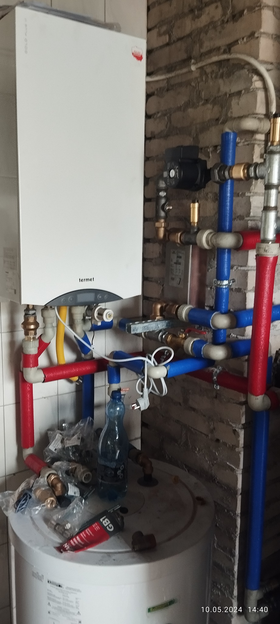 Kocioł gazowy Termet z podłączeniem instalacji grzewczej wykonanym z rur w kolorach czerwonym i niebieskim, widoczne akcesoria instalacyjne i butelka z wodą na zbiorniku.