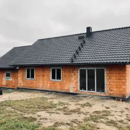 Surowy dom z czerwonej cegły z nowym, ciemnym dachem z dachówki ceramicznej i kominem, widok z zewnątrz na tle pochmurnego nieba.