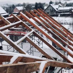 W trakcie budowy drewniana więźba dachowa pokryta śniegiem, na tle zimowego krajobrazu z ośnieżonymi domami i drzewami.