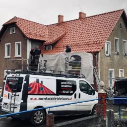 Ekipa dekarzy na rusztowaniu podczas układania nowej, czerwonej dachówki na dwupiętrowym domu z szarą elewacją, widoczny samochód firmowy z logo i danymi kontaktowymi.