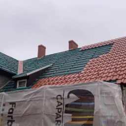 Częściowo wymienione czerwone dachówki na budynku, widoczna zielona konstrukcja pod dachówkami, rusztowanie z boku budynku osłonięte folią z logo Caparol.