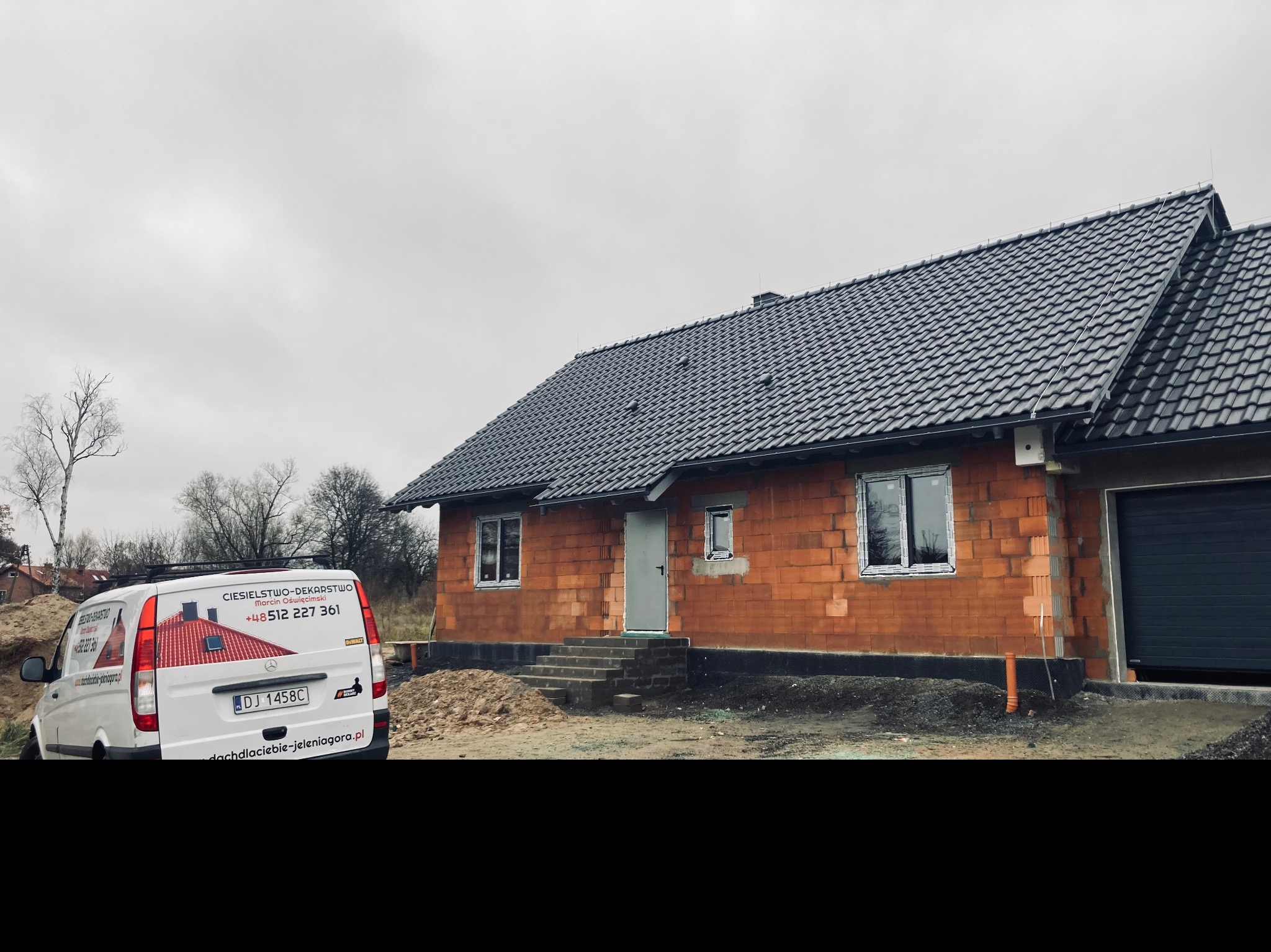 Dom w trakcie budowy z nowym, ciemnoszarym dachem z dachówki ceramicznej; na pierwszym planie samochód firmy dekarskiej z logo i numerem telefonu.
