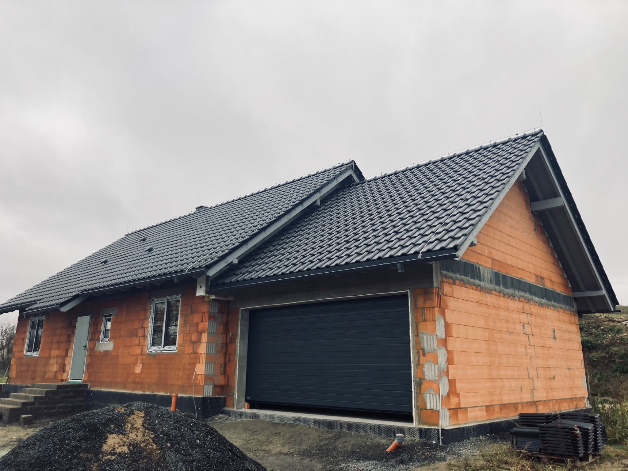 Surowy stan domu jednorodzinnego z szarą dachówką ceramiczną, widoczne niezadaszone wejście i brama garażowa, cegła dziurawka na ścianach zewnętrznych, przygotowana do dalszych prac wykończeniowych.