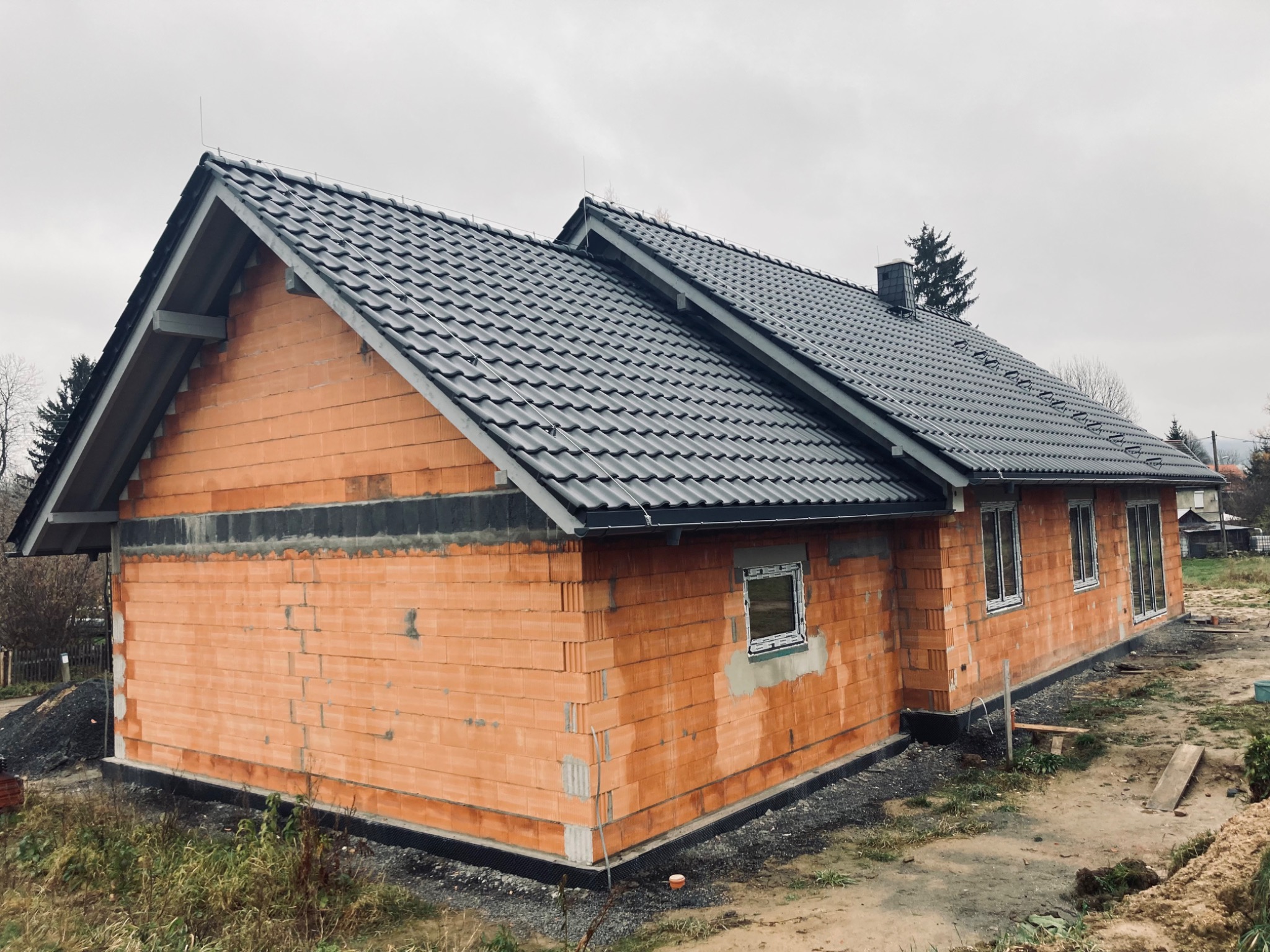 Niedokończony dom jednorodzinny z cegły ceramicznej, z zamontowanym ciemnoszarym dachem z dachówki ceramicznej i widocznym kominem. Okna częściowo zamontowane, teren wokół domu jeszcze...