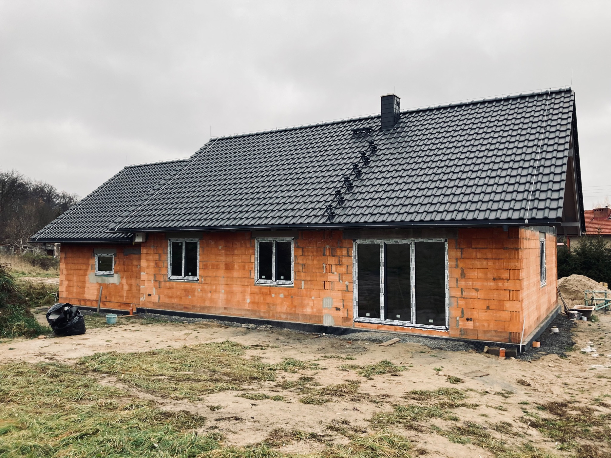 Surowy dom z czerwonej cegły z nowym, ciemnym dachem z dachówki ceramicznej i kominem, widok z zewnątrz na tle pochmurnego nieba.