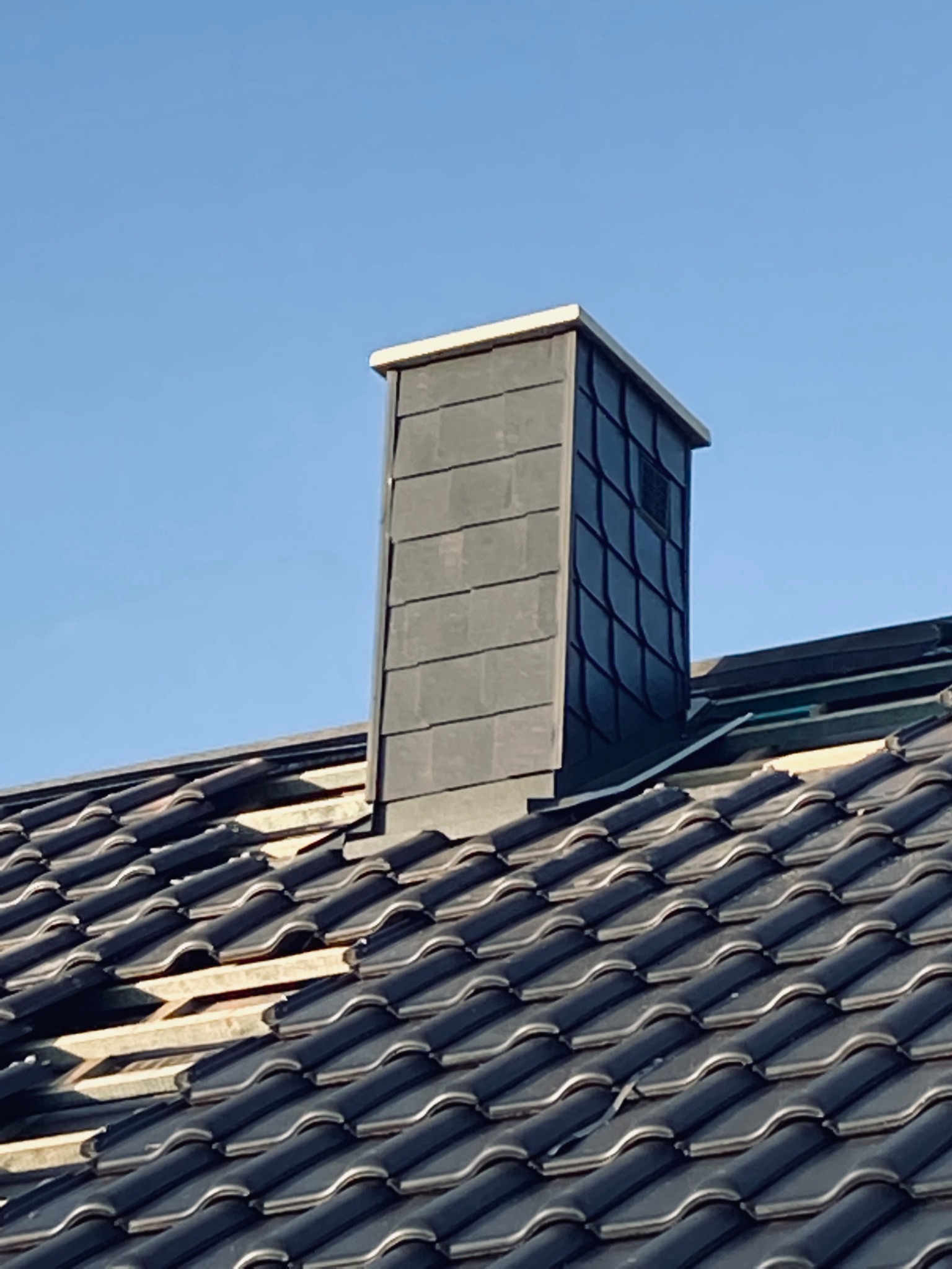 Nowoczesny komin obłożony szarymi płytkami na tle błękitnego nieba, widoczny na dachu z ciemnej dachówki ceramicznej, częściowo jeszcze bez pokrycia.