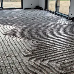 Rozłożone rury grzewcze na folii z siatką, przygotowane do zalania betonem w pomieszczeniu z dużymi oknami.