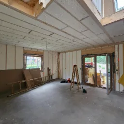 Wnętrze w trakcie prac wykończeniowych: ściany i sufit pokryte natryskową izolacją, widoczne drewniane elementy konstrukcyjne, okna, drzwi balkonowe, drabina, płyty budowlane i narzędzia na surowej...