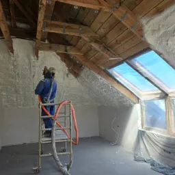 Pracownik w kombinezonie, stojąc na drabince, aplikuje pianę izolacyjną na poddaszu w Leśniewie, widoczne drewniane elementy konstrukcji dachu i okno dachowe zabezpieczone folią.