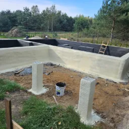 Fundament budynku po izolacji, pomalowany na czarno wewnątrz, z nałożoną warstwą jasnej pianki izolacyjnej z zewnątrz; widoczne dwa słupki również pokryte pianką, oraz wiadro po farbie na ziemi.