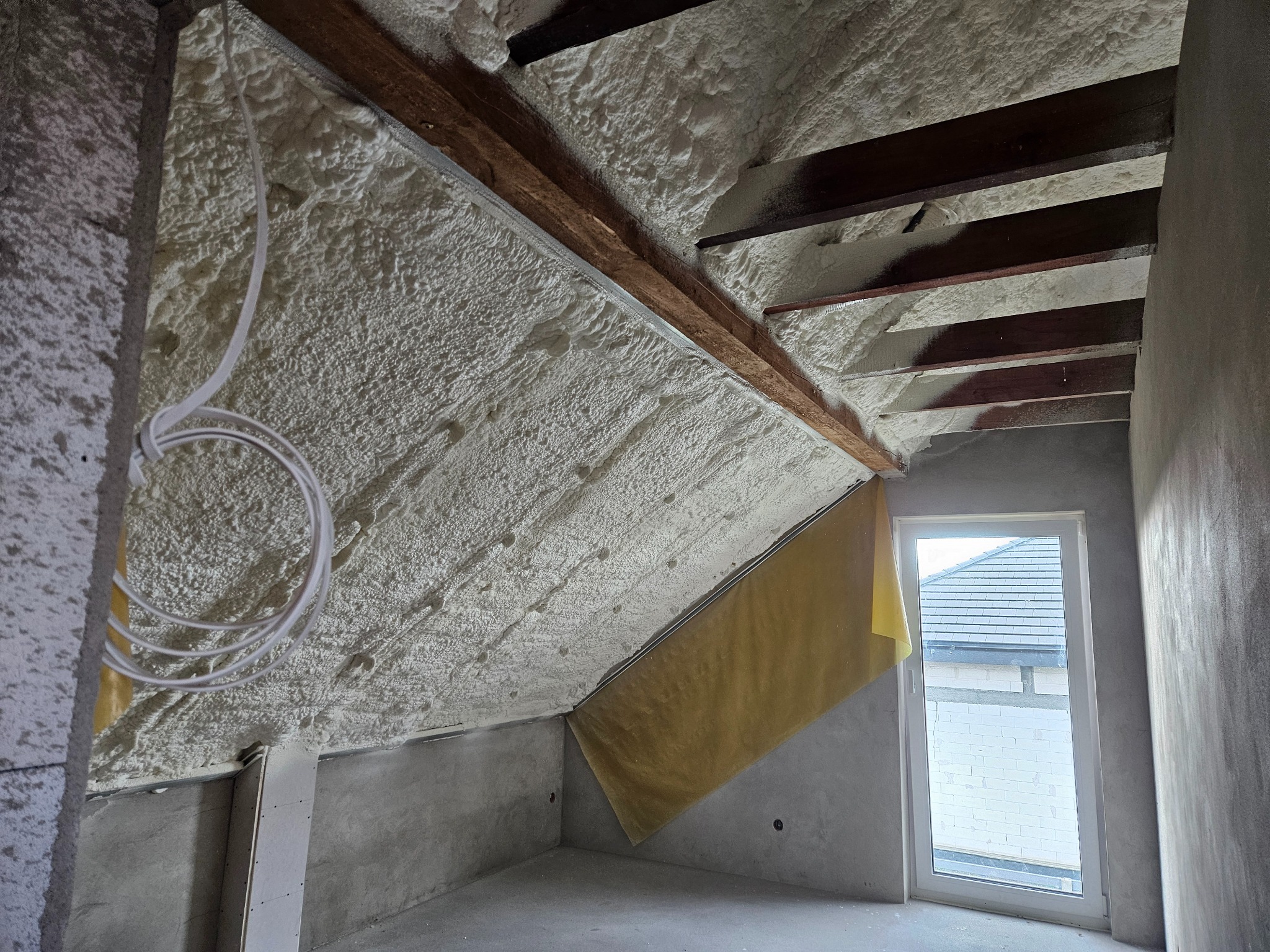 Poddasze z drewnianymi belkami po ociepleniu pianką, widoczne kable elektryczne i fragment okna z widokiem na dach sąsiedniego budynku.