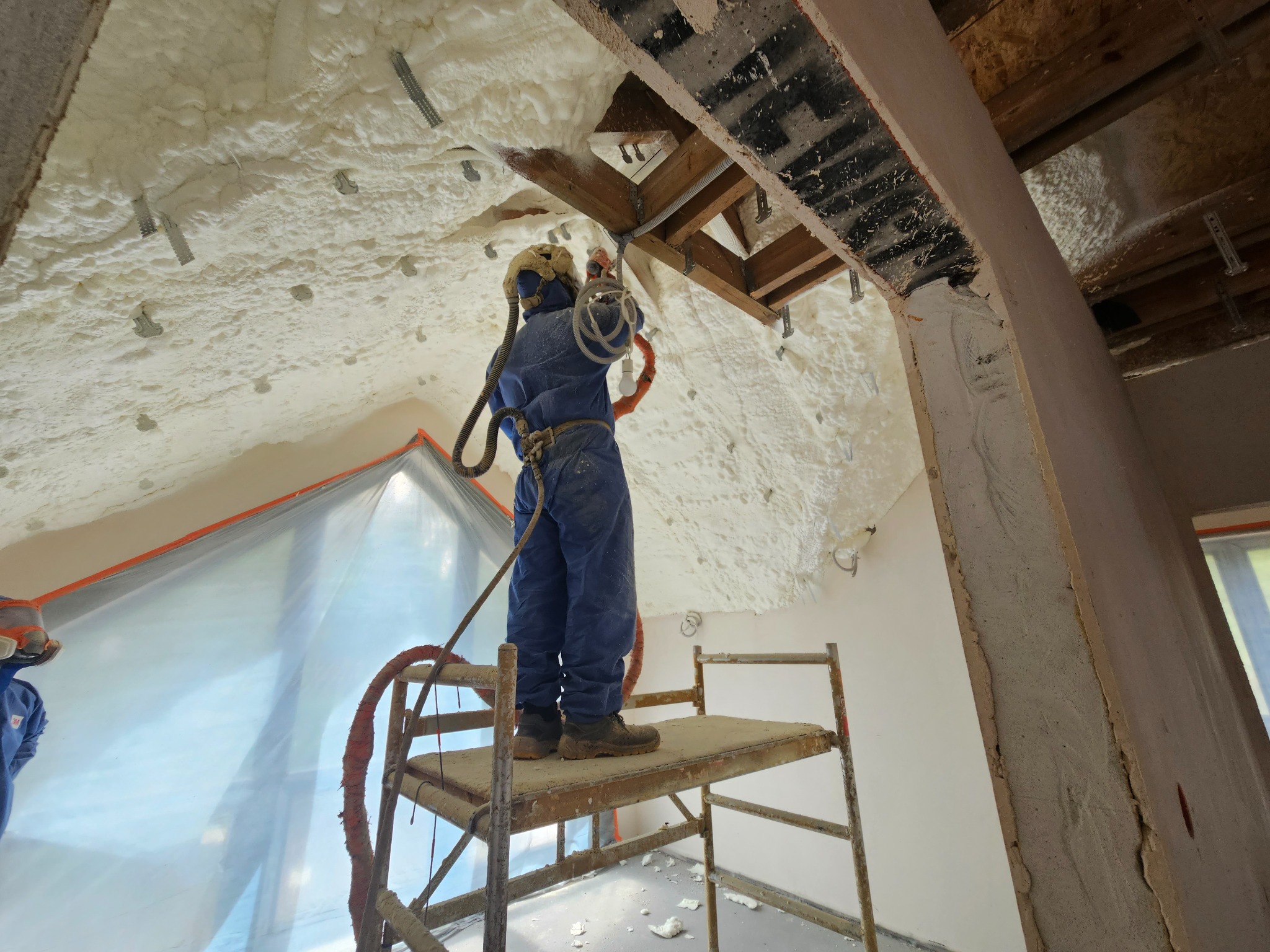 Pracownik w kombinezonie ochronnym na rusztowaniu aplikuje piankę izolacyjną na poddaszu, widoczne drewniane elementy konstrukcyjne i metalowe mocowania w pianie.
